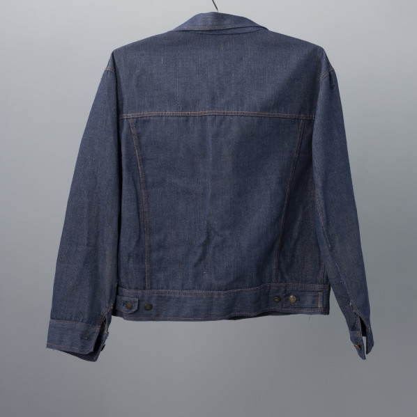 kurtka męska firmy ODRA - Ujęcie z tyłu; Krótka, granatowa kurtka męska z materiału dżinsowego. Na całej kurtce widoczne szycie pomarańczową nitką. Każdy guzik posiada wypukłą głowę gryfa zwróconą w lewa stronę oraz na otoku napis ODRA SZCZECIN.