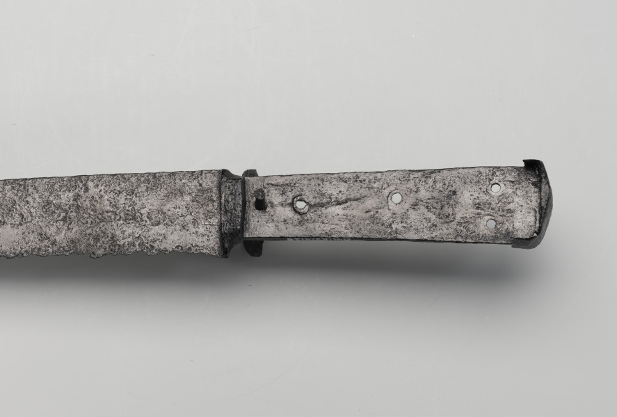 kord - Ujęcie 3/4 z dołu; Głownia kordu jest prosta, jednosieczna, o sztychu decentrycznym. Trzon z jednostronnym wyżłobionym wgłębieniem ma cztery otwory do mocowania okładzin rękojeści, których brak. Głowica jest w formie kapturka, czyli cienkiej stalowej blaszki wygiętej w kierunku ostrza.