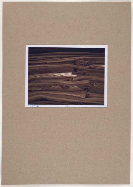 fotografia - Barwna odbitka fotograficzna w stonowanych kolorach przedstawiająca stos poskładanych tkanin leżących jedna na drugiej. Brak widocznych zakończeń tkanin wskazuje, że jest to kadr większej całości. Fotografia utrzymana jest w tonacji brązowo-beżowej z jasną plamą świetlną widoczną w centralnej części. Tkaniny leżą warstwami, płasko, w niektórych miejscach widoczne jest zawinięcie lub miejsca złożenia którejś z nich. W dwóch miejscach (w tym jednym z jaśniejszy oświetleniem), widoczny jest brzeg ozdobiony haftem i mereżką. Odbitka fotograficzna została wykonana z wąską, białą ramką.