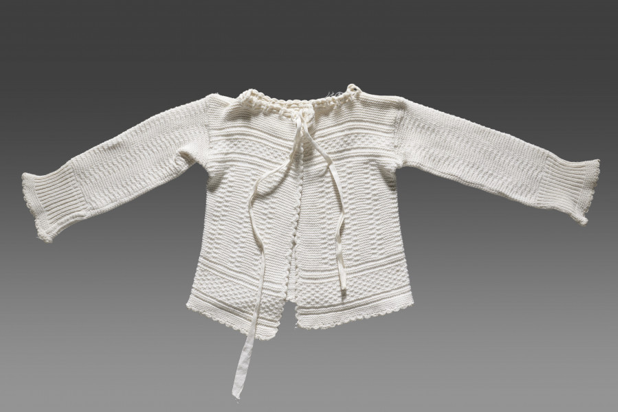 Kaftanik dziecięcy w kolorze écru. Ścieg układa się w geometryczny wzór. W centralnej części obiektu widoczny wzór kwadratów układających się w równoległe rzędy biegnące z góry do dołu. Na piesi oraz w dolnej części (około 1/3 wysokości od dolnej krawędzi) wzór układa się w dwa równoległe poziome pasy. Pomiędzy nimi ścieg jest równomierny (bez podziału na rzędy). Dolna krawędź wykończona falbanką. Kaftanik wiązany pod szyją na taśmę w tym samym kolorze. Rękawy zakończone ściągaczem.