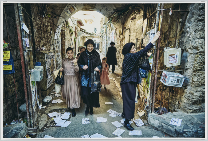 scena rodzajowa - Na pierwszym planie poziomego, barwnego kadru fotograficznego wydać dwie uśmiechające się dojrzałe kobiety oraz nastolatkę. Kobiety ubrane są w odświętne, zabudowane stroje z przewagą czerni, na głowach mają czarne nakrycia. Nastolatka ubrana jest w odświętna długa sukienkę w kolorze tzw. brudnego różu. Tuż za nimi idzie grupa dziewczynek (prawie niewidoczna, gdyż schowana za plecami idących na przedzie). Cała grupa idzie ulicą, przechodzi przez kamienne, bardzo zaśmiecone przejście, w którym umocowana jest otwarta na oścież metalowa brama. Jedna z kobiet dotyka wyciągniętą dłonią podłużnego, jasnego przedmiotu znajdującego się na odrzwiach bramy.