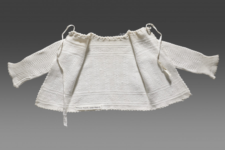 Kaftanik dziecięcy w kolorze écru. Ścieg układa się w geometryczny wzór. W centralnej części obiektu widoczny wzór kwadratów układających się w równoległe rzędy biegnące z góry do dołu. Na piesi oraz w dolnej części (około 1/3 wysokości od dolnej krawędzi) wzór układa się w dwa równoległe poziome pasy. Pomiędzy nimi ścieg jest równomierny (bez podziału na rzędy). Dolna krawędź wykończona falbanką. Kaftanik wiązany pod szyją na taśmę w tym samym kolorze. Rękawy zakończone ściągaczem.