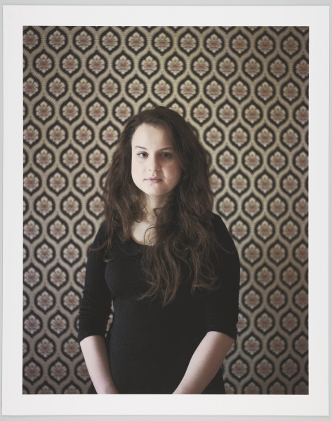portret fotograficzny - Fotografia barwna skadrowana pionowo, przedstawiająca półpostaciowy portret młodej kobiety. Portretowana dziewczyna o ciemnych, długich, falujących włosach, puszczonych luźno na ramiona, ubrana w ciemną prostą bluzkę z długimi rękawami podciągniętymi pod łokcie, patrzy prosto w stronę widza. Kobieta stoi we wnętrzu na tle ściany pokrytej tapetą o bardzo ozdobnym, symetrycznym, floralno-geometrycznym wzorze.