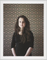 portret fotograficzny - Fotografia barwna skadrowana pionowo, przedstawiająca półpostaciowy portret młodej kobiety. Portretowana dziewczyna o ciemnych, długich, falujących włosach, puszczonych luźno na ramiona, ubrana w ciemną prostą bluzkę z długimi rękawami podciągniętymi pod łokcie, patrzy prosto w stronę widza. Kobieta stoi we wnętrzu na tle ściany pokrytej tapetą o bardzo ozdobnym, symetrycznym, floralno-geometrycznym wzorze.