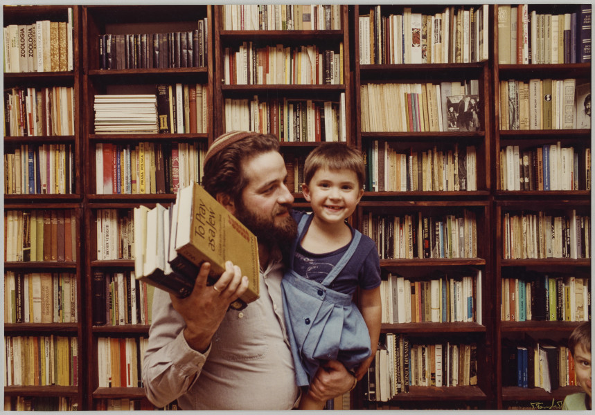 fotografia - Zdjęcie przedstawia mężczyznę z małą dziewczynka (córeczką) na ręku stojącego na tle regału z książkami (wypełniającego całe tło). Mężczyzna ma na sobie jasną koszulę i jarmułkę na głowie. Dziewczynka jest ubrana w niebieską sukienkę oraz ciemną koszulkę. Twarz mężczyzny jest zwrócona w stronę dziecka. Dziewczyna patrzy w obiektyw i się uśmiecha. W drugim ręku mężczyzna trzyma kilka książek.