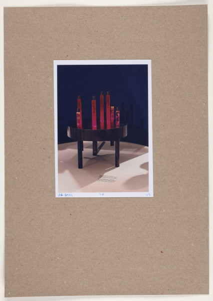 fotografia - Barwna odbitka fotograficzna przedstawiająca ciemny stół o masywnym blacie stojący na trzech walcowatych nogach. Do stołu zamontowane zostały kolejne dwie nogi, które są krótsze, zatem nie stanowią podpór. Na blatu stołu widocznych jest siedem wysokich, cylindrycznych elementów o różnej wysokości, ustawionych nieregularnie. Są to walce zakończone połyskliwymi, ciemnymi nasadkami, z przeźroczystego materiału o częściowo transparentnym wypełnieniu w nasyconych barwach (czerwono-pomarańczowych-różowych). W środku widoczne są pęcherze powietrza. Cała instalacja stoi we wnętrzu na tle ciemnej ściany i na białej podłodze, na której widoczny jest kilkuwersowy czarny napis umieszczony przed obiektem. Odbitka fotograficzna została wykonana z wąską, białą ramką.