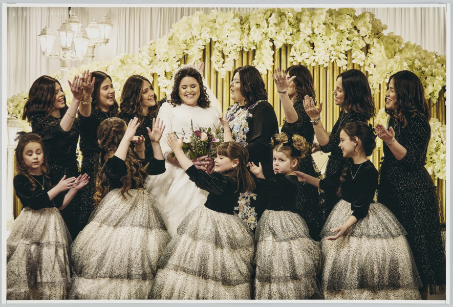 scena rodzajowa - Poziomy, barwny kadr fotograficzny przedstawiający pozowaną scenę ślubną. Na tle jasnej kotary ozdobionej girlandami jasnych, sztucznych kwiatów, białych lamp stoi grupa uśmiechniętych kobiet i dziewczynek w dwóch rzędach. W wyższym rzędzie po środku znajduje się panna młoda w białej sukni i z białym welonem po jej lewej i prawej stronie stoi 7 młodych kobiet w ciemnych sukienkach i z rozpuszczonymi ciemnymi, długimi włosami (3 po lewej i 4 po prawej) które są zwrócone w jej stronę. Poniżej stoi 5 dziewczynek w identycznych, odświętnych sukienkach o czarnej górze i tiulowej czarno-białej spódnicy. Zarówno kobiety, jak i dziewczynki maja podniesione ręce w geście prezentacji panny młodej.