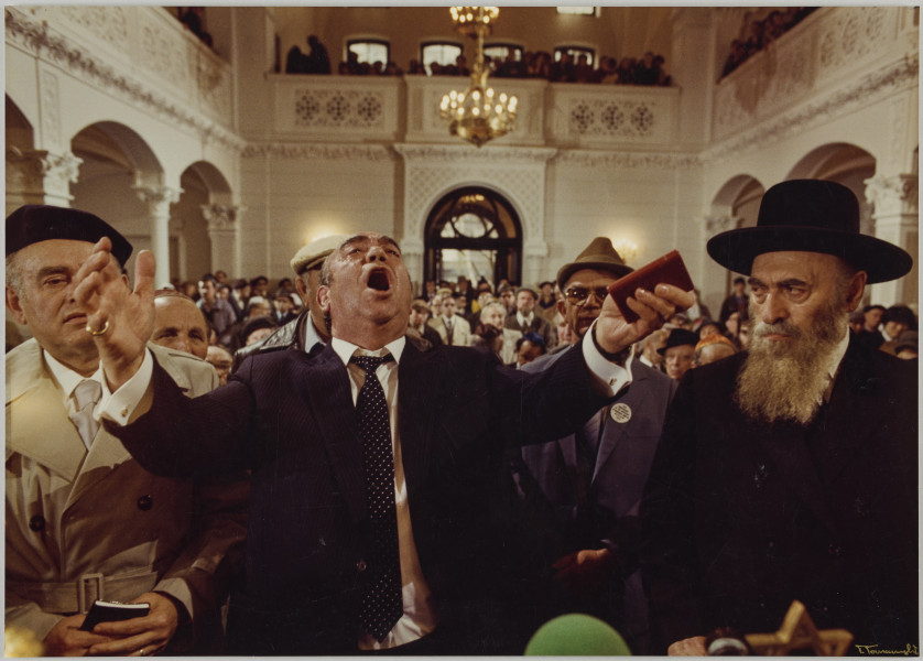 fotografia - Zdjęcie przedstawia wnętrze synagogi oraz modlących się w niej mężczyzn, którzy siedzą w kolejnych rzędach ławek. Po środku kompozycji, w pierwszej ławce widoczny mężczyzna z rozłożonymi szeroko rękami i otwartymi ustami. Ma na sobie garnitur, białą koszulę i granatowy krawat w drobne kropki. Na ławce obok niego siedzi dwóch mężczyzn (po lewej i prawej stronie). Mężczyzna po prawej ma na sobie kapelusz z szerokim rondem oraz garnitur i białą koszulę. Ma długą siwą brodę. Mężczyzna po lewej stronie ma na sobie białą koszulę, jasny krawat oraz beżowy płaszcz. Na głowie ma czarny beret. Za nimi widać rzędy ławek, w których siedzą pozostali mężczyźni. 