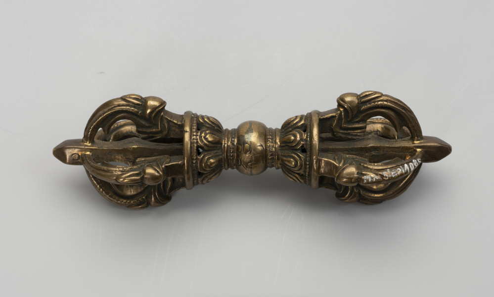 wadżra - ujęcie z przodu; Mały, wykonany z brązu, przypominający berło przedmiot w kształcie kokardy lub ósemki. W jego centrum znajduje się kula - uchwyt berła. Po jej obu stronach znajdują się wyrastające w przeciwnych kierunkach stożkowe zgrubienia stylizowane na kwiaty lotosu z ośmioma płatkami. Od każdego z nich odchodzą cztery, łukowate ramiona zbiegające się wokół centralnej szpicy przebiegającej przez całą oś obiektu. Ramiona bogato zdobione w formie ozdoby przypominającej podwinięty liść przedstawiającej wystylizowaną głową potwora zwanego makarą. Na kuli wygrawerowano napis, być może w piśmie tybetańskim.