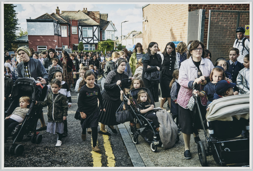 scena rodzajowa - Poziomy, barwny kadr fotograficzny przedstawiający dużą grupę, w której skład wchodzą kobiet, i dziewczęta. Trzy spośród nich, na pierwszym planie, pchają wózki z małymi dziećmi. Grupa zajmuje całą szerokość chodnika i ulicy pomiędzy niewysokimi miejskimi budynkami - idą w kierunku widza.