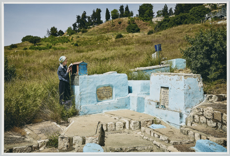scena rodzajowa - Poziomy, barwny kadr fotograficzny przedstawiający, w centralnej części kadru, kobietę ubraną w granatową bluzkę z długim rękawem oraz czarna spódnicę oraz chustkę zawiązana na głowie, czytającą książkę. Kobieta stoi obok niskich betonowych form architektonicznych z hebrajskimi inskrypcjami, pomalowanych na kolor jasnoniebieski. Obok są kamienno-betonowe płyty ułożone kaskadowo. Zabudowania znajdują się u stóp rozległego wzgórza, porośniętego łąką oraz niskimi krzewami. U góry kadru widoczne jest jednolite niebieskie niebo.