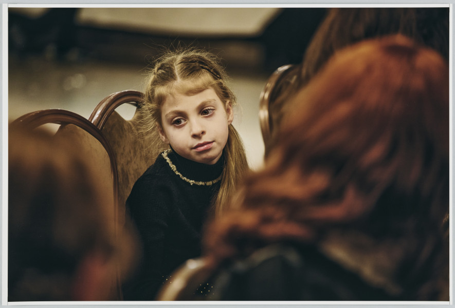 scena rodzajowa - Poziomy, barwny kadr fotograficzny przedstawiający portret małej dziewczynki. W centralnej części kadru siedzi na krześle mała dziewczynka o jasnych, długich, spiętych w kucyk włosach i ciemnych oczach, ubrana w czarny golf, na który ma założony sznurek jasnych koralików. Na pierwszym planie widoczne są sylwetki 3 postaci o długich kasztanowych włosach (ich zarysy są bardzo nie ostre).