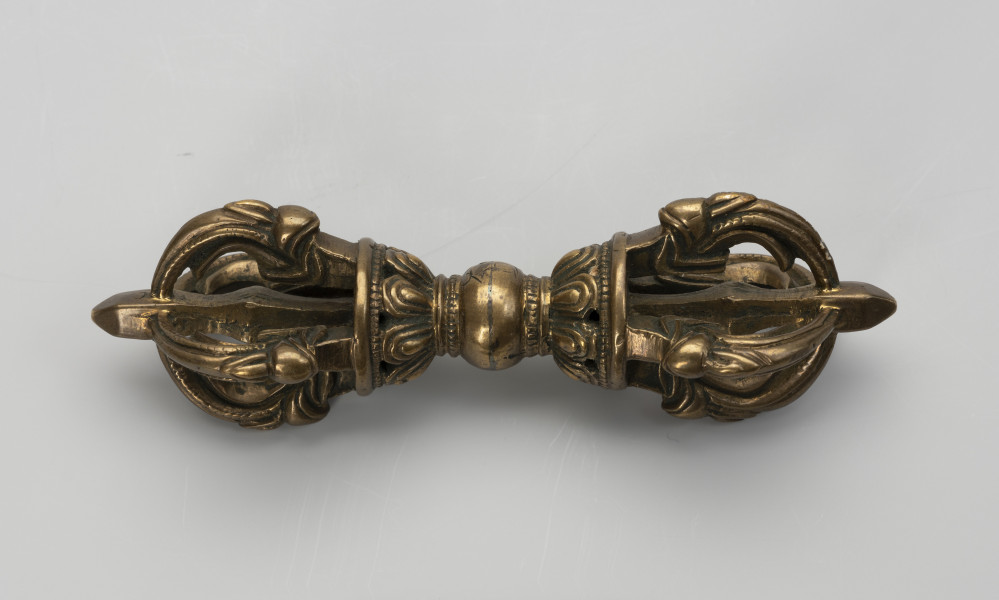 wadżra - ujęcie z przodu; Mały, wykonany z brązu, przypominający berło przedmiot w kształcie kokardy lub ósemki. W jego centrum znajduje się kula - uchwyt berła. Po jej obu stronach znajdują się wyrastające w przeciwnych kierunkach stożkowe zgrubienia stylizowane na kwiaty lotosu z ośmioma płatkami. Od każdego z nich odchodzą cztery, łukowate ramiona zbiegające się wokół centralnej szpicy przebiegającej przez całą oś obiektu. Ramiona bogato zdobione w formie ozdoby przypominającej podwinięty liść przedstawiającej wystylizowaną głową potwora zwanego makarą.