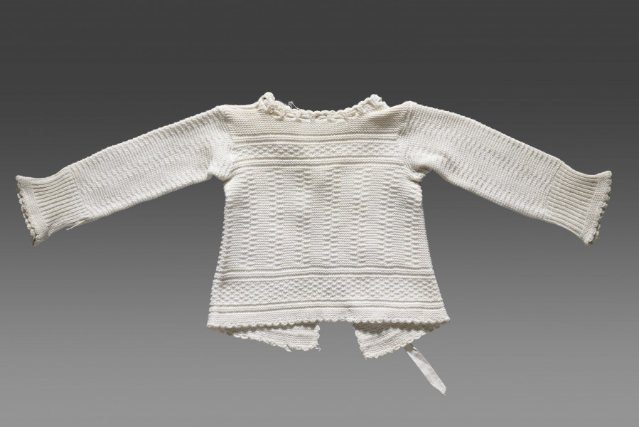 Kaftanik dziecięcy w kolorze écru. Ścieg układa się w geometryczny wzór. W centralnej części obiektu widoczny wzór kwadratów układających się w równoległe rzędy biegnące z góry do dołu. Na piesi oraz w dolnej części (około 1/3 wysokości od dolnej krawędzi) wzór układa się w dwa równoległe poziome pasy. Pomiędzy nimi ścieg jest równomierny (bez podziału na rzędy). Dolna krawędź wykończona falbanką. Kaftanik wiązany pod szyją na taśmę w tym samym kolorze. Rękawy zakończone ściągaczem.