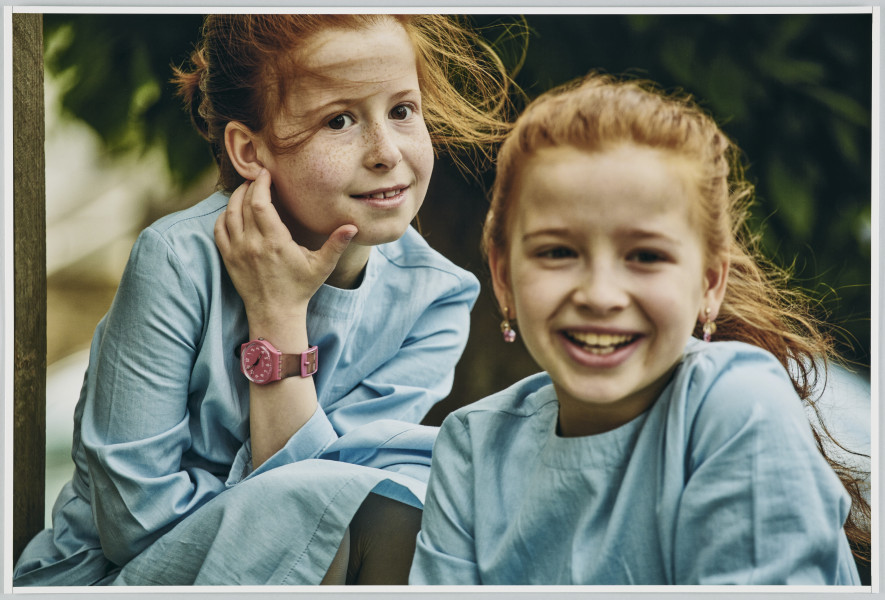 scena rodzajowa - Poziomy, barwny kadr fotograficzny przedstawiający dwie uśmiechnięte dziewczynki o rudych, długich włosach rozwiewanych przez wiatr. Dzieci siedzą blisko siebie, jedna na pierwszym planie, druga tuż za nią. Obie są ubrane w jednakowe niebieskie sukienki. Dziewczynka z tyłu ma na lewej ręce różowy zegarek.