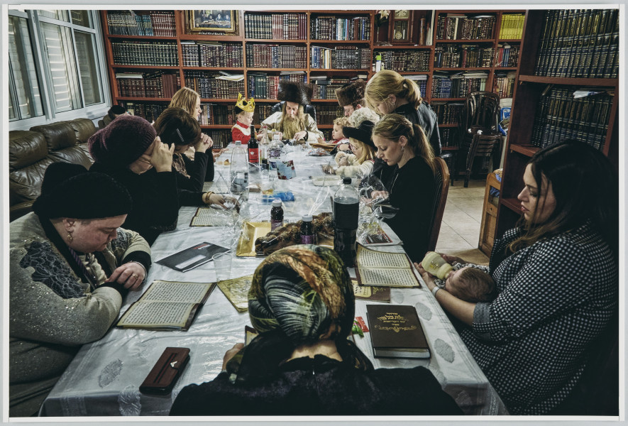 scena rodzajowa - Poziomy, barwny kadr fotograficzny przedstawiający grupę kobiet i dziewczynek siedzących przy długim stole. U szczytu stołu siedzi także dwóch mężczyzn z brodami, długimi pasmami włosów zakrywających uszy (pejsami) oraz w dużych futrzanych czapach w kształcie walca. Obok mężczyzn stoi dwoje małych dzieci. Zebrani pogrążeni są w czytaniu tekstów leżących przed każdą z osób, ich pozy wskazują na skupienie. Jedna z kobiet czyta i karmi butelką niemowlę leżące na jej kolanach. Stół stoi po środku pomieszczenia, którego ściany są zastawione w całości regałami, na których stoją książki w dekoracyjnych obwolutach (biblioteka?). Ścianę po lewej tronie kadru w całości wypełnia zasłonięte okno. Na stole nakrytym obrusem i przezroczystą ceratą stoją w części opróżnione butelki po napojach oraz rozpakowane folie i puste talerze. w pomieszczeniu pełnym regałów z książkami.