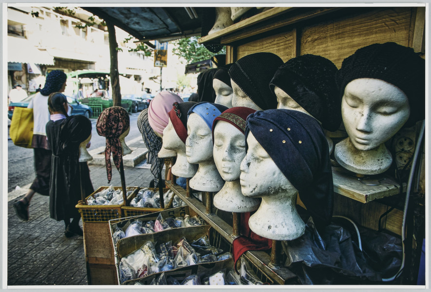 scena rodzajowa - Poziomy, barwny kadr fotograficzny, na którym widoczna jest miejska ulica. Na pierwszym planie, po prawej stronie i w centralnej części widoczny jest stragan z kilkunastoma białymi manekinami głów. Na nich umieszczone są różnorodne i różnobarwne damskie nakrycia głowy (chustki i czapki). Na drugim planie, po lewej stronie widać kobietę w chustce i spódnicy idącą z dziewczynką w sukience. W tle nieostre zarysy budynków stojących wzdłuż ulicy.
