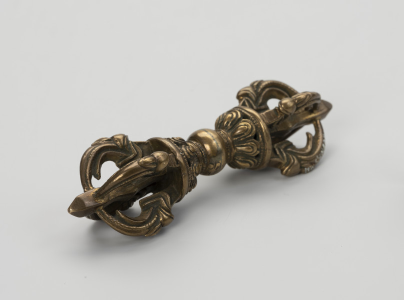 wadżra - ujęcie przodu ze skosu z lewej; Mały, wykonany z brązu, przypominający berło przedmiot w kształcie kokardy lub ósemki. W jego centrum znajduje się kula - uchwyt berła. Po jej obu stronach znajdują się wyrastające w przeciwnych kierunkach stożkowe zgrubienia stylizowane na kwiaty lotosu z ośmioma płatkami. Od każdego z nich odchodzą cztery, łukowate ramiona zbiegające się wokół centralnej szpicy przebiegającej przez całą oś obiektu. Ramiona bogato zdobione w formie ozdoby przypominającej podwinięty liść przedstawiającej wystylizowaną głową potwora zwanego makarą.