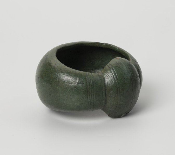 kopia bransolety nerkowatej z Kolkowa - Ujęcie przodu ze skosu z lewej; Bransoleta o szerokiej obręczy (5,9 cm), silnie wypukła. Jej brzegi są mocno zagięte do środka, co daje C-kształtny przekrój poprzeczny. Oba końce obręczy łączy duże zgrubienie, tzw. nerka. Końce i zgrubienie nerkowate zostały ozdobione pasmami zwielokrotnionych linii (3 pasma przypadają na zgrubienie, a 4 pasma na końce obręczy), które biegną poprzecznie w stosunku do obręczy. Pomiędzy pasmami na zgrubieniu nerkowatym znajduje się tzw. ornament oczkowy (współśrodkowe podwójne okręgi z centralnie usytuowaną kropką-wgłębieniem). Bransoleta ma zielony kolor. Brzeg nerki ma w jednym miejscu zamarkowane uszkodzenie, wzorowane na oryginale bransolety.