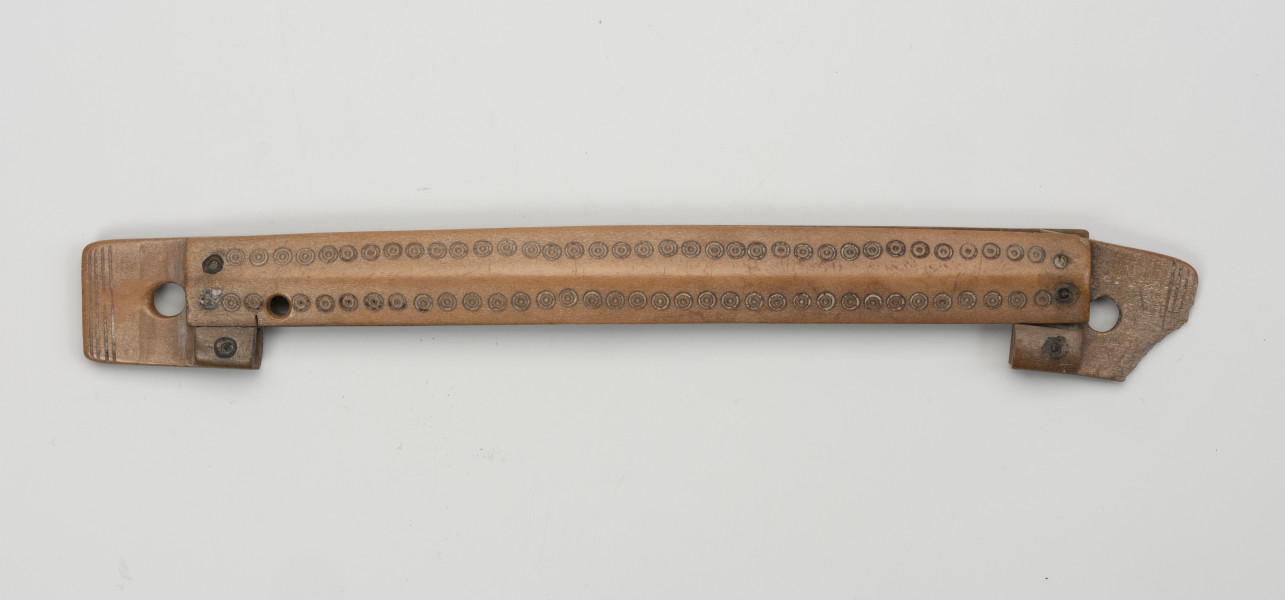 pochewka grzebienia - Ujęcie z przodu; Obiekt zbudowany został z dwóch okładzin o kształtach wydłużonych prostokątów o prostych bokach, okładziny, ułożone równolegle względem siebie, umocowane są do dwóch bocznych płytek za pomocą nitów wykonanych ze stopu miedzi, po dwa nity na każdą płytkę. Boczne płytki częściowo wchodzą w odstęp pomiędzy okładzinami, wyznaczając w ten sposób szerokość przestrzeni na grzebień, a częściowo wystają poza ich końce. Jedna ich krawędź położona jest w tej samej linii co krawędź okładzin, a druga nieco wystaje. Na tych wystających odcinkach umocowano dodatkowo krótkie fragmenty kolejnych okładzin, zrównanych z jednej strony z okładzinami głównymi, a z drugiej z krawędziami wewnętrznymi płytek. Płytki posiadają koliste otwory do umocowania grzebienia w pochewce, a ich zakończenia zdobione są czterema nacięciami biegnącymi przez całą szerokość. Okładziny główne ozdobiono przy krawędziach dwoma rzędami kolistych oczek z zaznaczonymi środkami. Całość została wypolerowana.