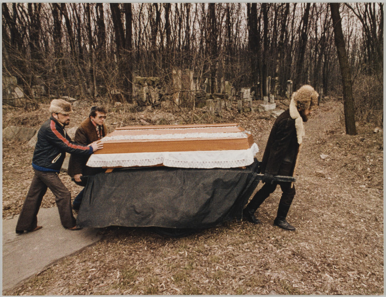 fotografia - Zdjęcie przedstawia trzech mężczyzn niosących trumnę na metalowych noszach. Jeden z mężczyzn znajduje się z przodu trumny (po prawej stronie kadru), dwaj z tyłu (po lewej stronie kadru). Nosze są przykryte ciemną tkaniną sięgającą niemal ziemi. Trumna jest wykonana z drewna. Spod wieka wystają krawędzie białej tkaniny wykończonej koronką. Samo wieko także zostało ozdobione taśmami białej koronki przytwierdzonymi wzdłuż krawędzi. Mężczyzna idący z przodu trumny ma na sobie krótki kożuch oraz futrzaną czapkę. Jeden z mężczyzn idących z tyłu trumny trzyma uchwyty noszy. Drugi kładzie ręce na wieku. Mężczyzna trzymający nosze ma na sobie ciemną (brązową) kurtkę, okulary i jarmułkę na głowie. Mężczyzna dotykający wieka ma na sobie krótką sportową kurtkę z czerwonym lampasem biegnącym wzdłuż rękawa oraz futrzaną czapkę. W tle widać stare macewy oraz drzewa bez liści (zdjęcie wykonane jesienią lub w zimie). 