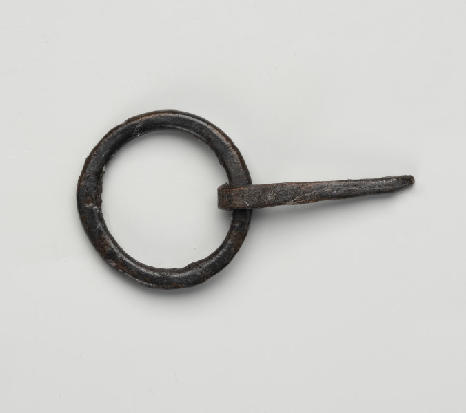 sprzączka do pasa - Ujęcie z dołu; Kolista sprzączka żelazna ze spłaszczonego drutu z ruchomym kolcem na ramie. Kolec przymocowano poprzez wygięcie tępego końca drutu i owiniecie go wokół obręczy sprzączki. Na powierzchni zachowane są pojedyncze włókna.