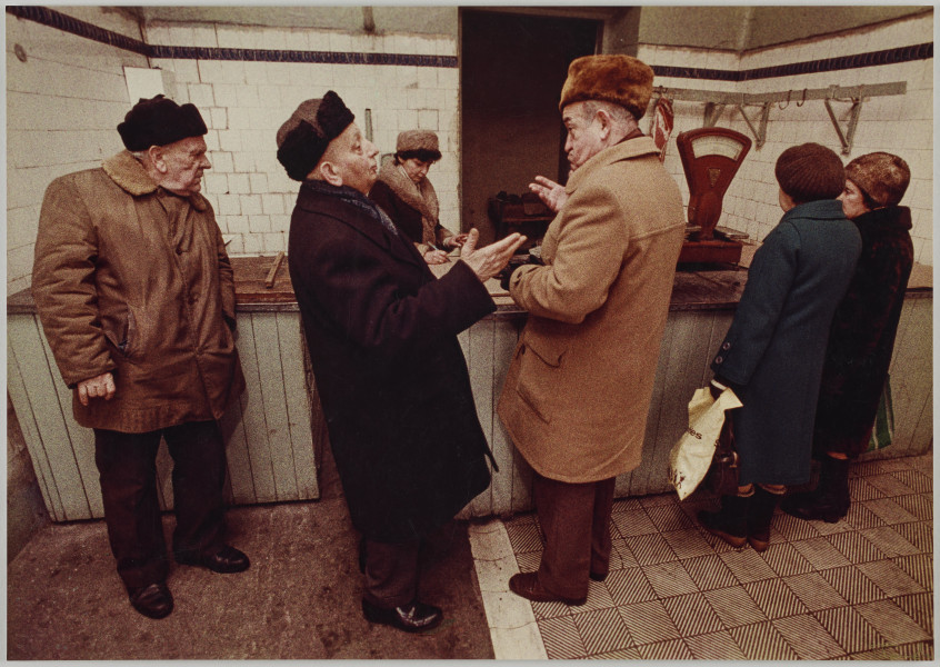 fotografia - Zdjęcie przedstawia grupę osób przy ladzie sklepowej. Po środku kadru widać dwóch rozmawiających ze sobą mężczyzn. Są zwróceni twarzami ku sobie. Gestykulują. Mają na sobie ciepłe płaszcze (płaszcz mężczyzny po lewej jest ciemny, po prawej jasny) oraz futrzane czapki. Po ich lewej stronie (na końcu kolejki) stoi starzy mężczyzna w kożuchu, ciemnej futrzanej czapce oraz ciemnych spodniach. Jego głowa jest zwrócona w stronę lady. Po prawej stronie rozmawiających mężczyzn (na początku kolejki; przy ladzie) stoją dwie kobiety w ciemnych płaszczach i czapkach. Ich twarze są zwrócone do lady. Stoją do niej przodem. Przed nimi (na ladzie) widać wagę sklepową. Za ladą stoi kobieta. Ma na siebie kożuch i futrzaną czapkę. Zapisuje coś w zeszycie, który leży przed nią. Ściany pomieszczenia wyłożone są białymi płytkami. Po prawej stronie widoczny przytwierdzany do ściany poprzeczny drążek, a na nim haki. Na jednym z haków ( po lewej stronie) wisi kawałek mięsa.