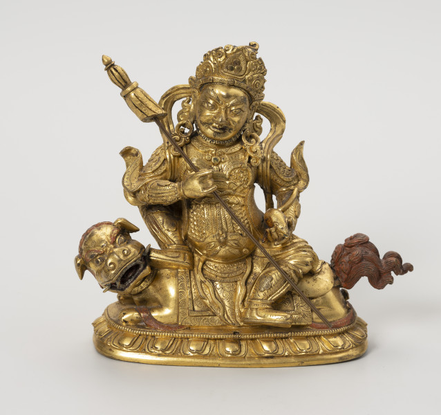 Wajśrawana (Dzambala) - ujęcie z przodu; Złota, męska postać siedząca bokiem, z lewą nogą opuszczoną w dół (poza lalitasana), na złotym, ryczącym lwie. W prawej ręce trzyma chorągiew zwycięstwa (osobny, wyciągany element), a w lewej zwierzątko: mangustę  (ichneumon), która wypluwa klejnot z pyszczka. Postać w gniewnej formie, szlachetnie ubrana: nosi diadem, liczne ozdoby z klejnotów, a jej strój przypomina zbroję wysokiej rangi dostojnika/króla. Lwa o bordowych brwiach, sierści na głowie i ogonie przedstawiono w pozycji wpółleżącej - waruje na ugiętych łapach, jest osiodłany, z przodu i z tyłu widoczna uprząż. Obie postacie ustawiono na lotosowej podstawie. W jej centralnym punkcie wydrążono niewielki otwór na szpic sztandaru. Posążek najprawdopodobniej przedstawia bóstwo bogactwa Wajśrawanę wraz z jego atrybutami.