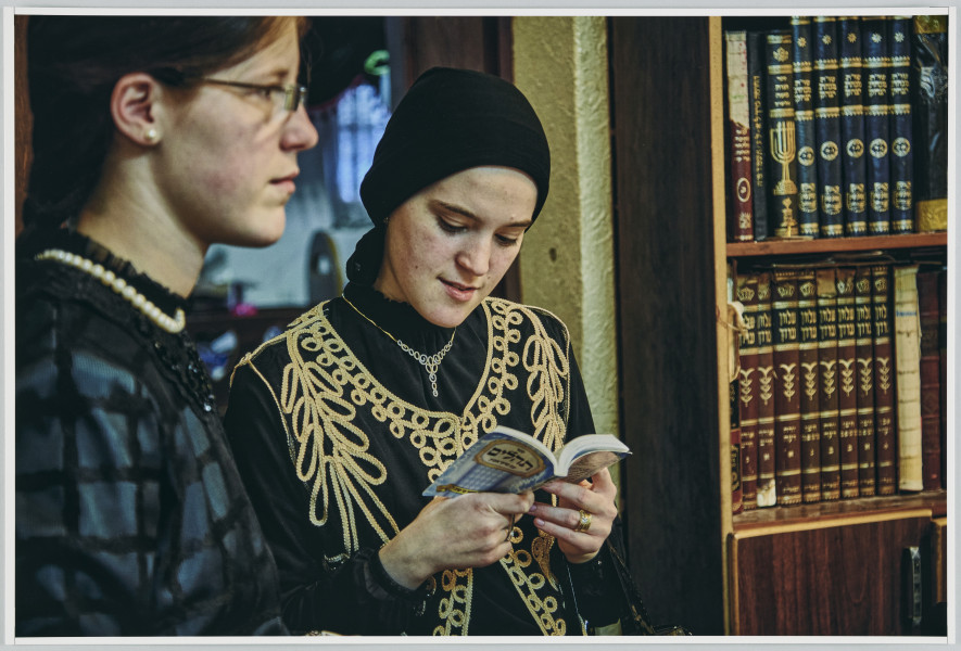 scena rodzajowa - Poziomy, barwny kadr fotograficzny przedstawiający dwie młode kobiety stojące we wnętrzu. Sylwetka jednej z nich, w okularach, w czarnej bluzce i krótkim sznurem białych pereł na szyi, widoczna z prawego profilu, wypełnia lewą stronę kadru. Dziewczyna patrzy przed siebie. Druga kobieta stoi koło niej. Jest ubrana w czarna bluzkę, czarną kamizelkę ozdobioną złotą wstążką w duże wzory, nosi na szyi srebrny niewielki naszyjnik oraz czarną chustę na głowie. W ręku trzyma książeczkę w języku hebrajskim i czyta z niej. Po prawej stronie widoczny jest ciemnobrązowy regał wypełniony ściśle książkami o ozdobnych grzbietach.