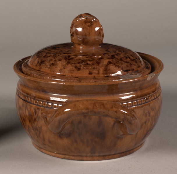 Miseczka okrągła z dwoma uchwytami i pokrywką. Pokrywka zakończona uchwytem w kształcie pąka kwiatu. Całość w kolorze wypalonej gliny