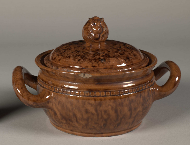 Miseczka okrągła z dwoma uchwytami i pokrywką. Pokrywka zakończona uchwytem w kształcie pąka kwiatu. Całość w kolorze wypalonej gliny