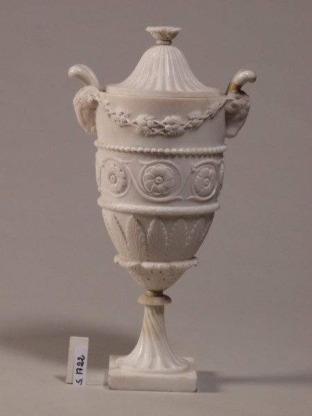 Wazon w stylu klasycystycznym z uchwytem w kształcie głów baranów. Pokrywka dzwonowata.