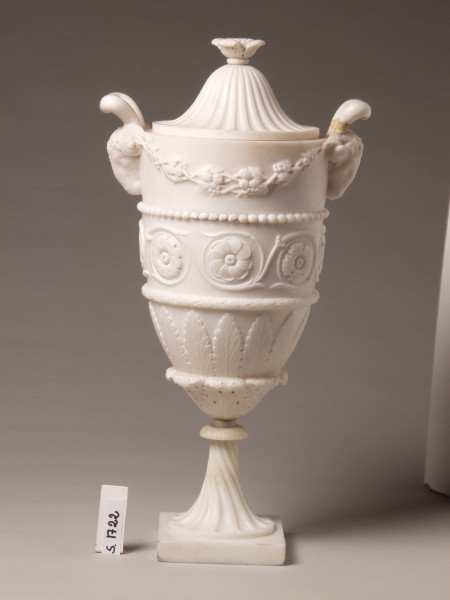 Wazon w stylu klasycystycznym z uchwytem w kształcie głów baranów. Pokrywka dzwonowata.