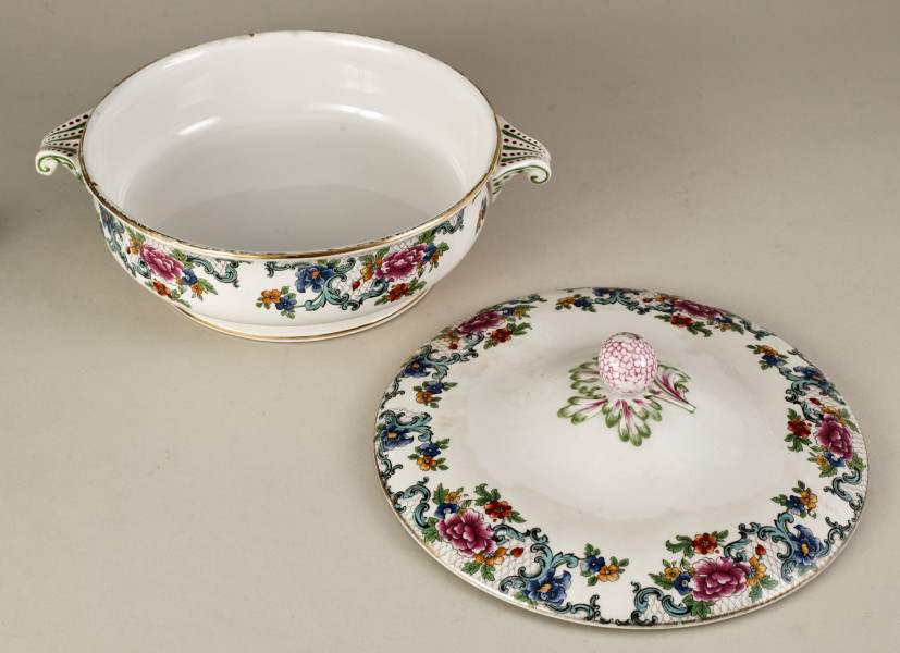 Półmiskowa waza z pokrywką, wykonana z białego fajansu, obiekt w kształcie cylindra. Naczynie ozdobione wielobarwną kalkomanią złożoną z asymetrycznych roślinnych kompozycji, wzory nawiązują do sztuki rokoko.