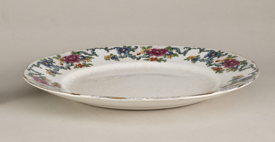 Stołowy, płytki talerzyk wykonany z białego fajansu. Zdobiony wielobarwną kalkomanią, złożoną z czterech jednakowych asymetrycznych roślinnych kompozycji