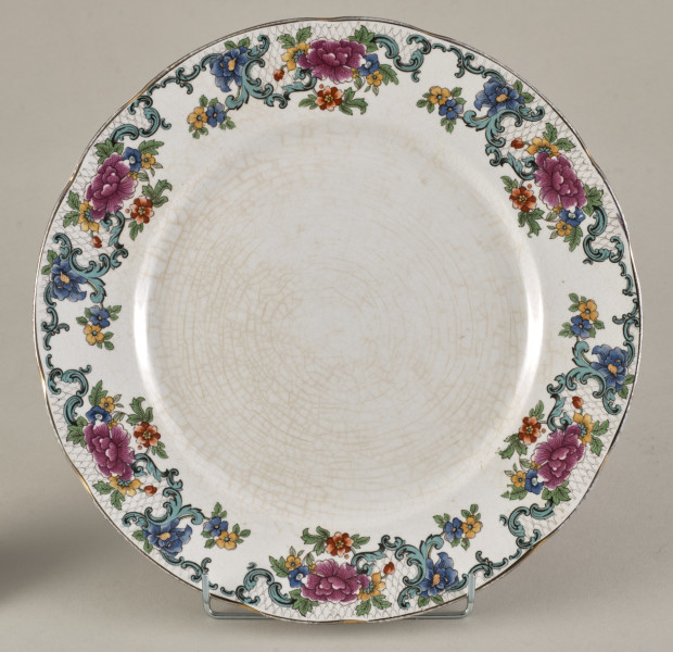 Stołowy, płytki talerzyk wykonany z białego fajansu. Zdobiony wielobarwną kalkomanią, złożoną z czterech jednakowych asymetrycznych roślinnych kompozycji